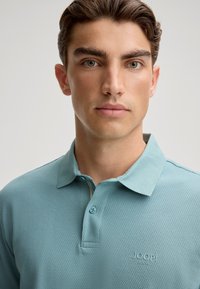 Polo bleu clair en tissu texturé avec un col classique, deux boutons et un petit logo embossé sur le côté gauche de la poitrine.