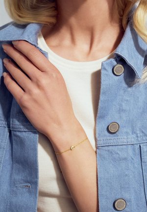 Guld kædearmbånd med en lille halvmånevedhæng, båret på en hånd. Modellen har en lys blå denimjakke og en hvid top på.