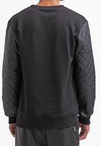 Persona che indossa una felpa nera con maniche in pelle trapuntata, vista di spalle, in piedi contro uno sfondo neutro.