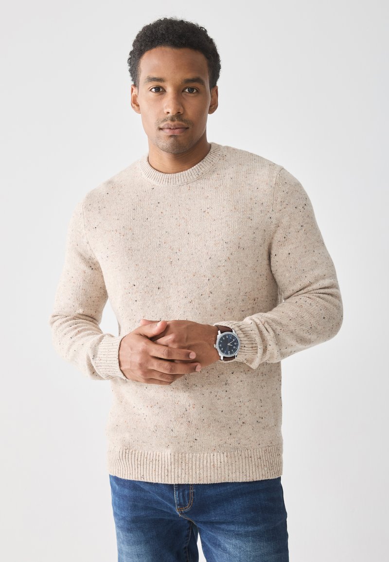 Beiger Strickpullover mit strukturierten Sprenkeln. Gerippter Rundhalsausschnitt und Bündchen. Kombiniert mit blauen Jeans und einer Uhr. Einfaches, lässiges Design.