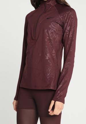 Vrouw die een bordeauxrode lange mouw top met een kwart-ritssluiting draagt, voorzien van subtiele bloemmotieven, gecombineerd met bijpassende bordeauxrode legging.