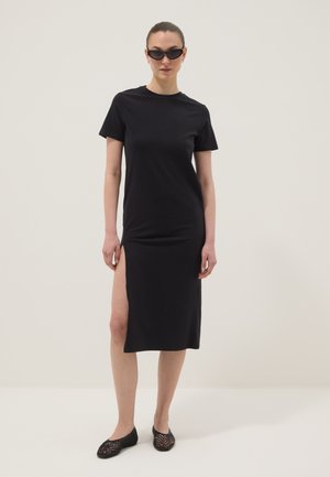 Frau trägt ein schwarzes Midi-Kleid mit kurzen Ärmeln und hohem Schlitz, schwarze gewebte flache Schuhe und dunkle Sonnenbrille, steht vor einem schlichten hellen Hintergrund.