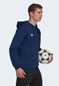 Marineblå hettegenser med kengurulomme, snor i hetta, og hvit Adidas-logo på brystet; holder en svart og hvit fotball.