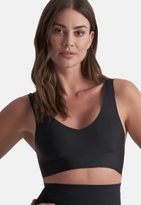 Kvinna med vågigt brunt hår som bär en svart sport-bh och högmidjade leggings, stående mot en enkel vit bakgrund.