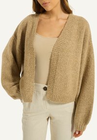 Beige gestrickte Strickjacke mit tiefem V-Ausschnitt, weiten Ärmeln und geripptem Saum. Kombiniert mit hellen Hosen für einen geschichteten Look.