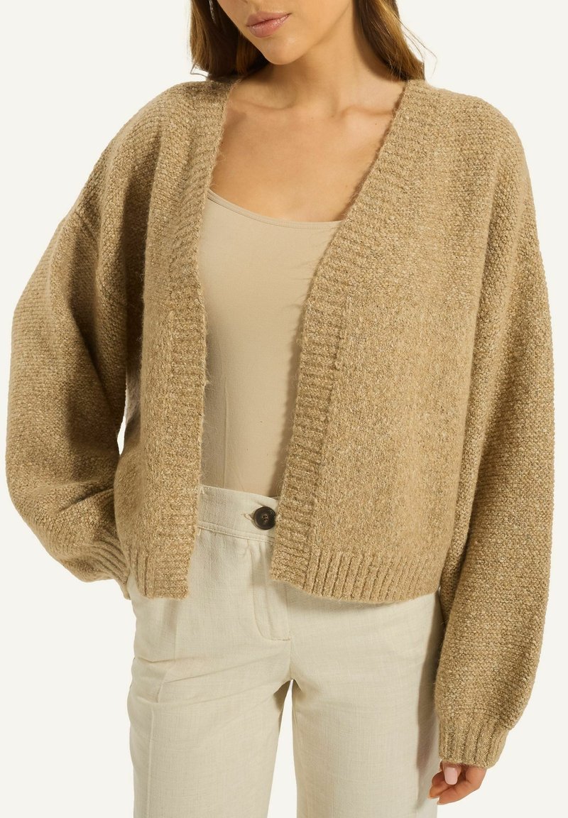 Beige gestrickte Strickjacke mit tiefem V-Ausschnitt, weiten Ärmeln und geripptem Saum. Kombiniert mit hellen Hosen für einen geschichteten Look.