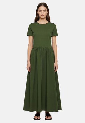 Donna che indossa un abito lungo verde oliva a maniche corte con vita arricciata e sandali neri aperti, in piedi davanti a uno sfondo semplice.