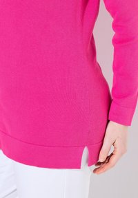 Fuchsia långärmad stickad topp med sidofickor, rundad nederkant och ribbade manschetter, ihop med vita byxor.