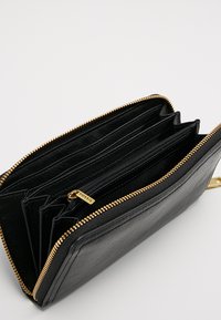 Portafoglio in pelle nera con zip dorata, dotato di più scomparti per carte e di un compartimento con zip per le monete. Superficie testurizzata con accentuazione del logo.
