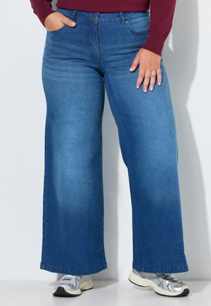 Person iført viddeblå jeans, sølvfarvede sneakers og en vinrød langærmet top med den ene hånd i lommen.
