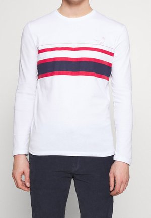 T-shirt bianco a maniche lunghe con strisce orizzontali rosse e blu navy, con una piccola grafica di un uccello sopra le strisce. Materiale in cotone.
