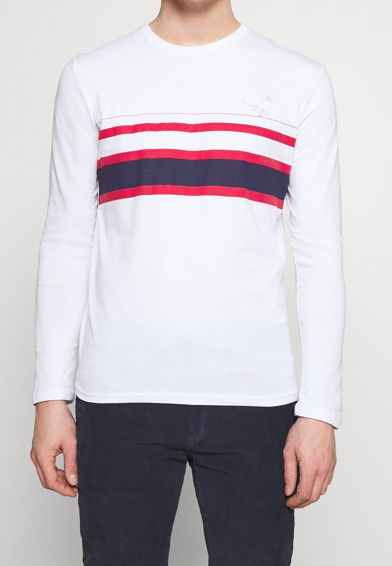 Langärmliges weißes T-Shirt mit rot-navy horizontalen Streifen, das ein kleines Vogelgrafik über den Streifen aufweist. Aus Baumwollmaterial.