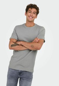 Camiseta gris de manga corta hecha de un tejido suave. Cuenta con un cuello redondo y un ajuste relajado. El modelo tiene tatuajes visibles en los brazos.
