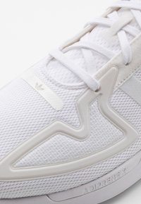 Scarpa sportiva bianca con tomaia in rete, dettagli in plastica testurizzata e logo "Adiprene X" sulla suola. Presenta lacci piatti e un design minimalista.