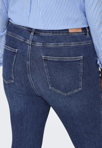 Jeans de denim azul oscuro con un ajuste slim, que cuentan con dos bolsillos traseros y costuras de contraste claras, combinados con una camisa a rayas azul y blanca.
