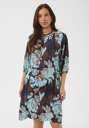Frau trägt ein lockeres, knielanges Kleid mit blau-braunem Blumenmuster und steht vor einem schlichten, hellen Hintergrund, nach vorne gerichtet.