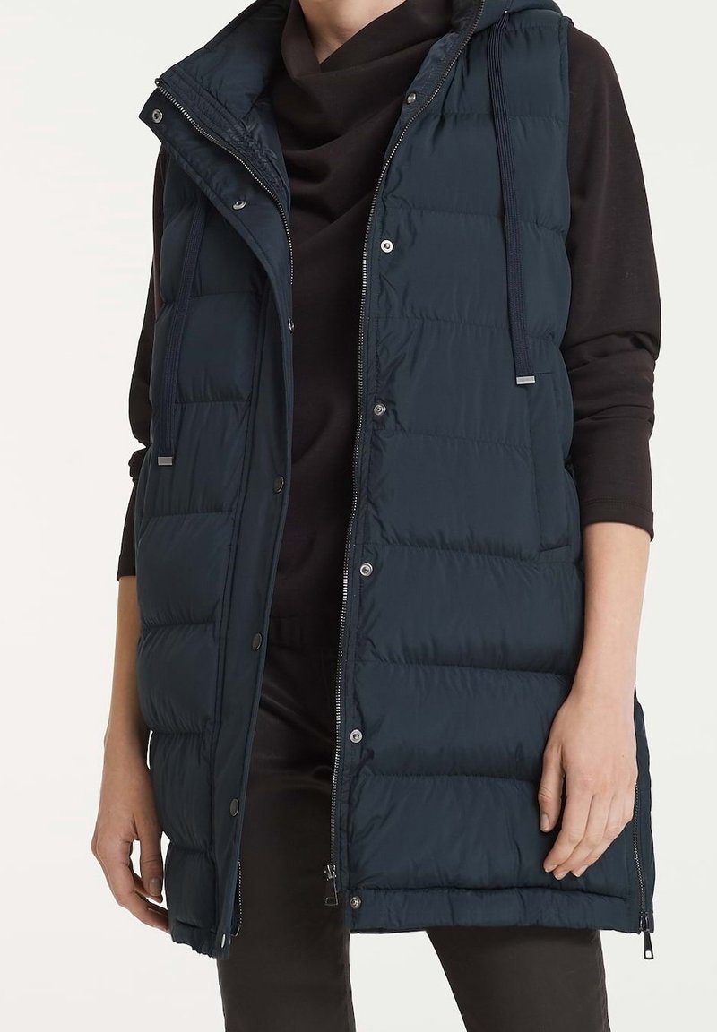 Femme portant un gilet matelassé sans manches bleu marine sur un haut noir à manches longues, les mains détendues le long du corps.