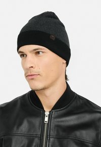 Junger Mann, der eine schwarz-graue Strickmütze und eine schwarze Lederjacke mit Reißverschluss vorne trägt und leicht zur Seite blickt.
