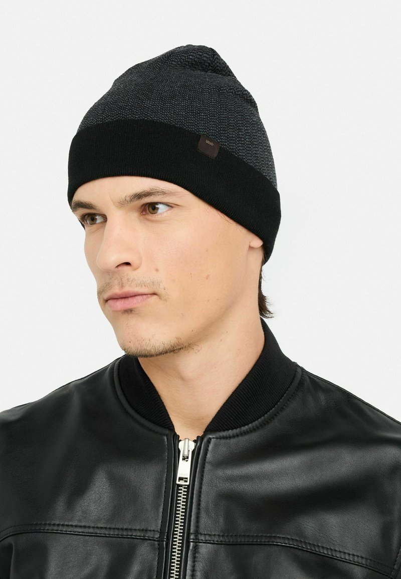 Junger Mann, der eine schwarz-graue Strickmütze und eine schwarze Lederjacke mit Reißverschluss vorne trägt und leicht zur Seite blickt.