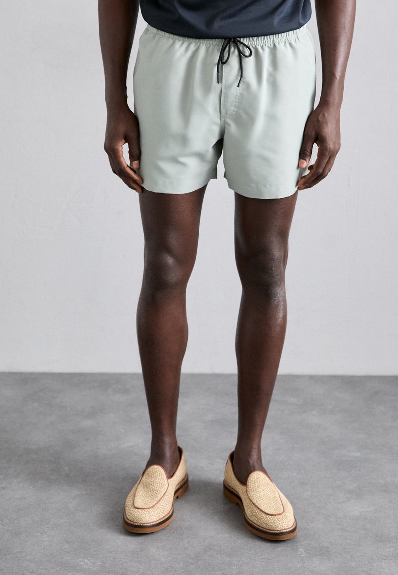 Shorts de bain vert clair avec une taille élastique et un cordon de serrage. Portés avec des chaussures beiges à enfiler, dotées d'une texture tissée et d'accents marron.