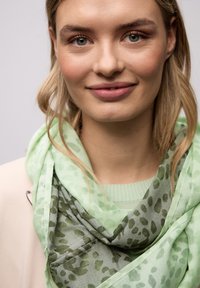 Jeune femme aux cheveux blond clair portant une écharpe verte à motifs et un pull clair, souriant doucement devant un fond neutre.