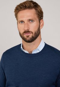 Suéter azul marino con cuello redondo, hecho de una tela suave. Llevado sobre una camisa de color azul claro con cuello, que presenta sutiles rayas horizontales.