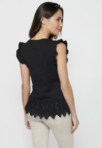 Haut noir à manches courtes avec des manches volantées et un ourlet festonné en œillets, fabriqué en tissu doux, associé à un pantalon beige clair.