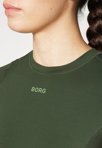 Žalia sportinė marškinėlių viršus su apvaliomis iškirptėmis, pagamintas iš lygios medžiagos. Priekyje yra kontrastingas šviesiai žalias "BORG" logotipas.