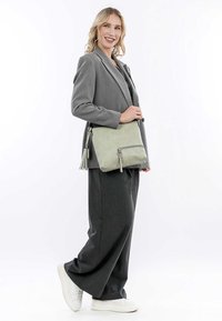 Svetle zelená kožená crossbody taška se zipem, detailem v podobě třásní a jemnou texturou, kterou nosí model v šedém obleku a bílých botách.