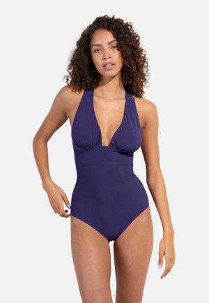 Donna con capelli ricci che indossa un costume intero viola scuro con collo halter e scollatura profonda, in piedi davanti a uno sfondo semplice.