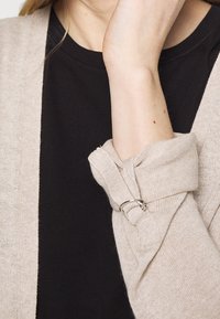 Béžový pletený svetr s volným střihem a vyhrnutými rukávy upevněnými stříbrným kroužkem. Pod ním černé tričko s hladkou texturou.