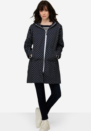 Impermeabile blu con pois, con zip frontale, cappuccio e due tasche laterali. Realizzato in materiale impermeabile. Abbinato a jeans scuri e sneaker bianche.