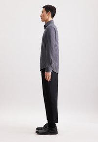 Camicia grigia a maniche lunghe con motivo, abbinata a pantaloni neri su misura, caratterizzati da una vestibilità slim, polsini bottonati e una leggera coda sul retro.
