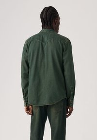 Πράσινο πουκάμισο από τζιν με μακριά μανίκια και μυτερό γιακά. Διαθέτει καμπυλωτό τελείωμα, κλείσιμο με κουμπιά και λεπτομέρειες στο πίσω μέρος.