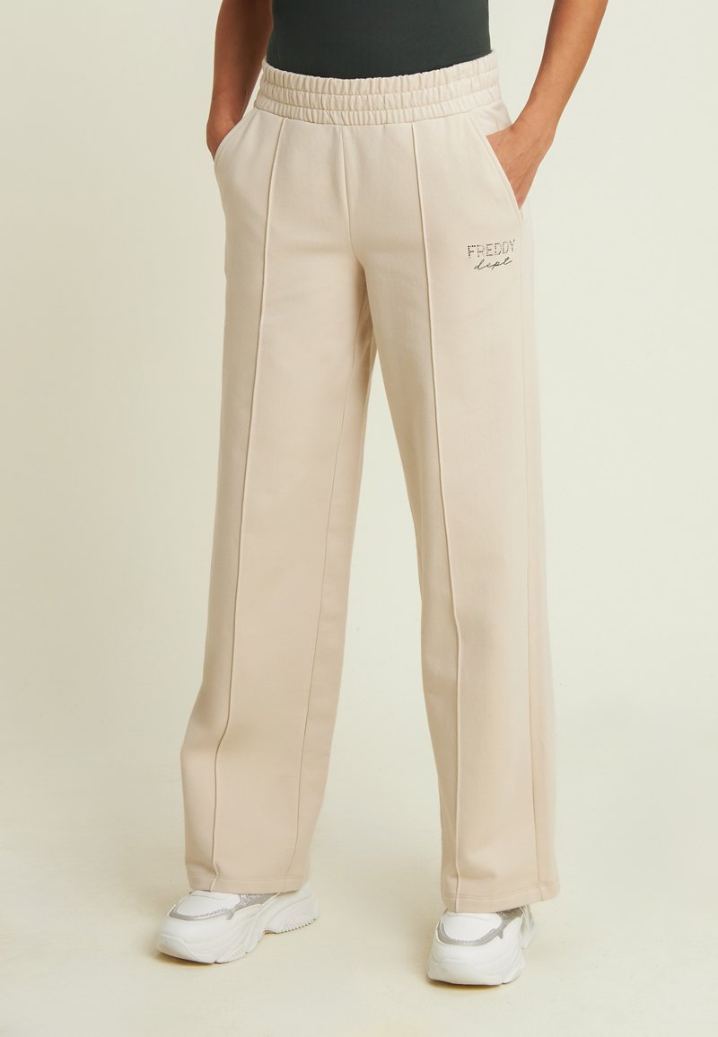 Pantalones de chándal de pierna ancha en color beige, con cintura elástica, bolsillos laterales y detalle del logo "Freddy". La tela parece suave y lisa.