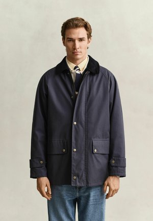 Junger Mann trägt eine dunkelblaue Jacke über einem hellen Hemd, Krawatte und Jeans und steht vor einem einfarbigen beigen Hintergrund.