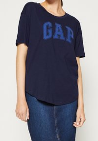 Námořnická modrá bavlněná tričko s kulatým výstřihem a krátkými rukávy. Na přední straně je umístěno logo "GAP" v modré barvě. Zakřivený lem.