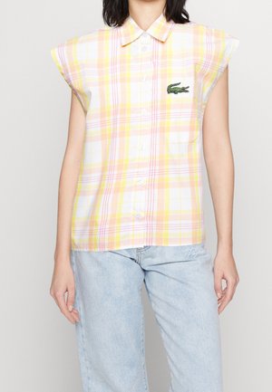 Kvinde iført en gul og pink ternet ærmeløs skjorte med en lille grøn krokodillelogo og lyseblå jeans.