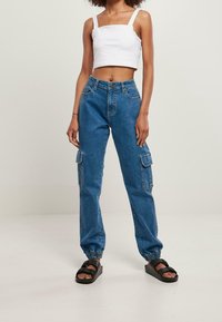 Pantalones cargo de mezclilla azul con bolsillos laterales, puños en los tobillos ajustados y una textura resistente. Usados con un top corto blanco y sandalias negras.