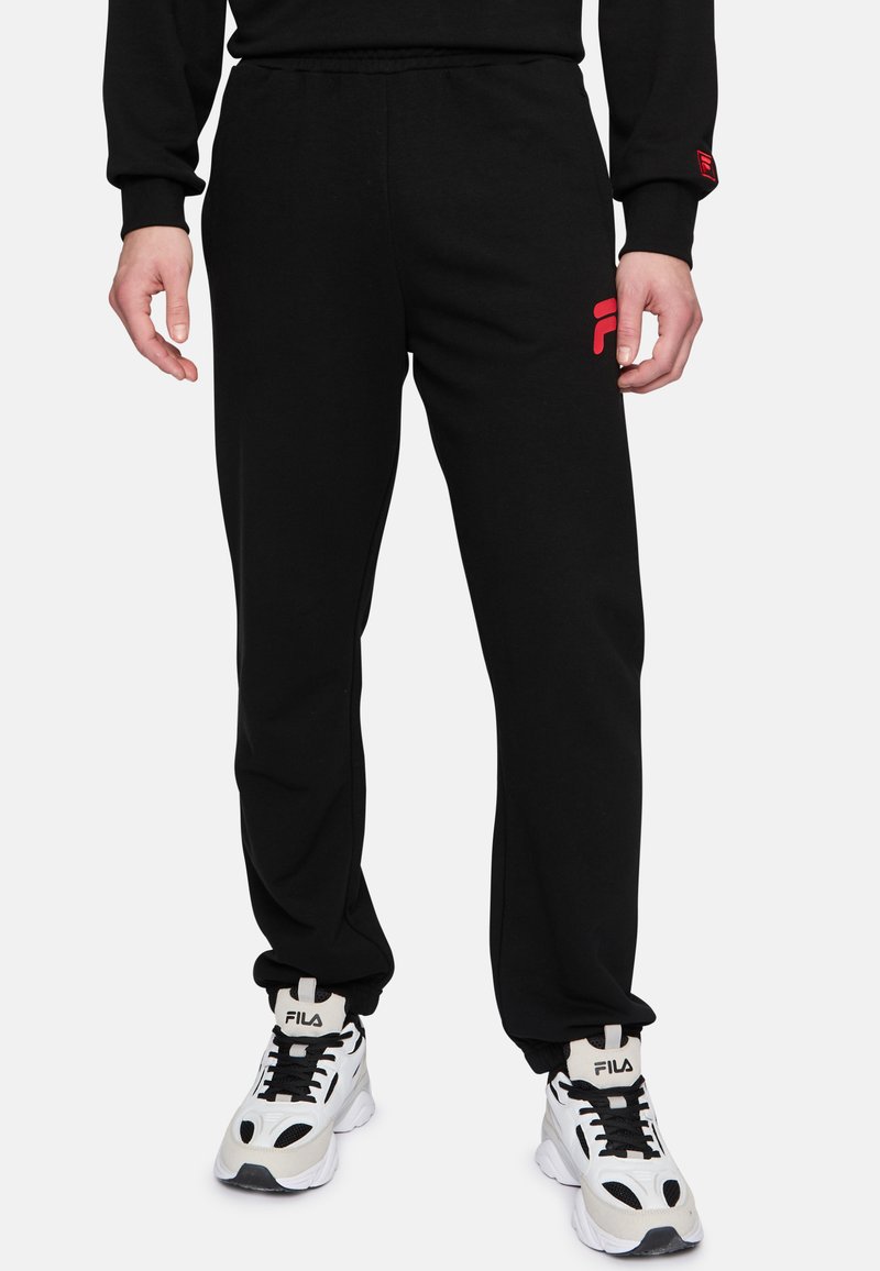 Zwarte trainingsbroek met elastische tailleband, zijzakken en een rood Fila-logo op de linkerdij. Draagt met witte sneakers met zwarte accenten.