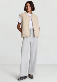 Beige ärmellose Weste mit Stehkragen, kombiniert mit einem weißen T-Shirt und grauen Hosen mit weitem Bein. Schwarze Schuhe mit runden Akzenten.