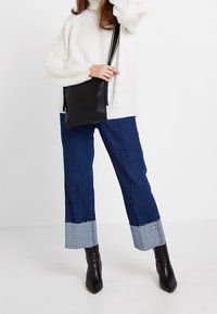 Biały gruby sweter z dzianiny, ciemnoniebieskie szerokie dżinsy z jasnoniebieskimi mankietami z denimu oraz czarne botki za kostkę. Czarna torba na ramię.