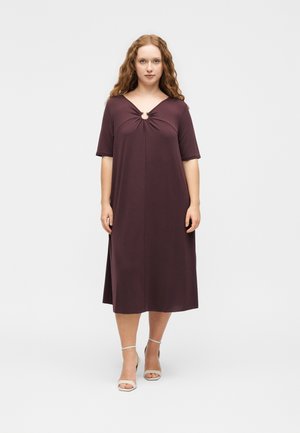 Γυναίκα με μακριά σγουρά μαλλιά που φοράει μωβ σκούρο φόρεμα μέχρι το γόνατο με λεπτομέρεια δαχτυλιδιού στο ντεκολτέ και λευκά ψηλά παπούτσια με λουράκι στον αστράγαλο, στέκεται όρθια.