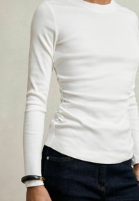 Cămașă albă cu mâneci lungi, cu detalii pliate pe lateral, asortată cu blugi de denim închis. Are un decolteu rotund și o croială ajustată.
