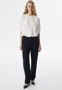 Witte blouse met korte mouwen en een zak, gecombineerd met donkerblauwe, op maat gemaakte broek. Model draagt zwarte, puntige ballerina's. Strak, minimalistisch ontwerp.