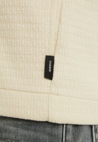 Tissu beige texturé avec une étiquette de vêtement noire portant l'inscription « CHASIN' » cousue près de la couture au-dessus d'un jean en denim gris.