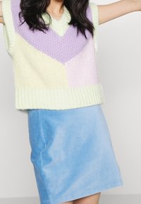 Gebreide mouwloze trui in pastelgroen, paars en geel met een V-hals en geometrisch patroon, gecombineerd met een blauwe corduroyen minirok.