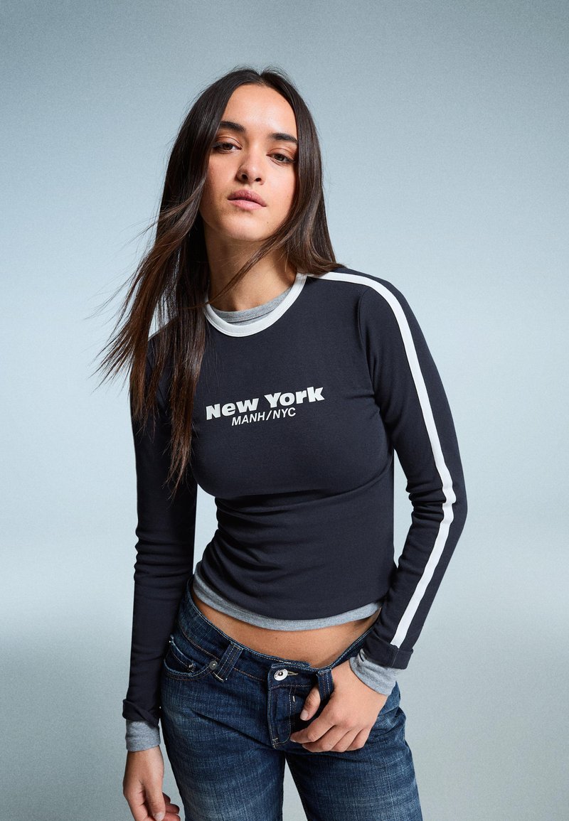 Fekete, hosszú ujjú ing fehér csíkokkal és "New York" grafikával, kék denim farmerrel párosítva. Szürke részletek a gallérnál és az ujjaknál.