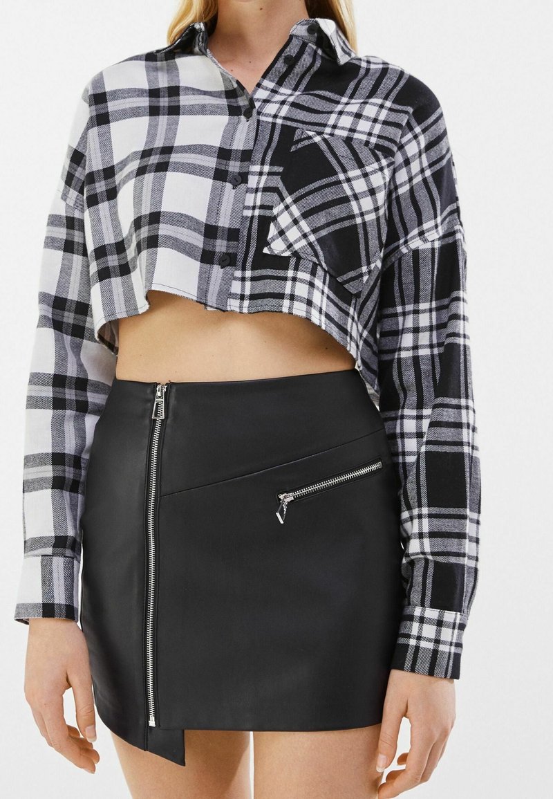 Femme portant une chemise courte à carreaux noir et blanc et une mini-jupe noire avec des détails de zips à l'avant et sur les côtés.