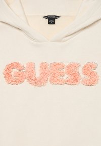 Kremljana dukserica s pastelnim ružičastim cvjetnim teksturiranim slovima koja ispisuju "GUESS" obrubljena malim srebrnim perlicama.